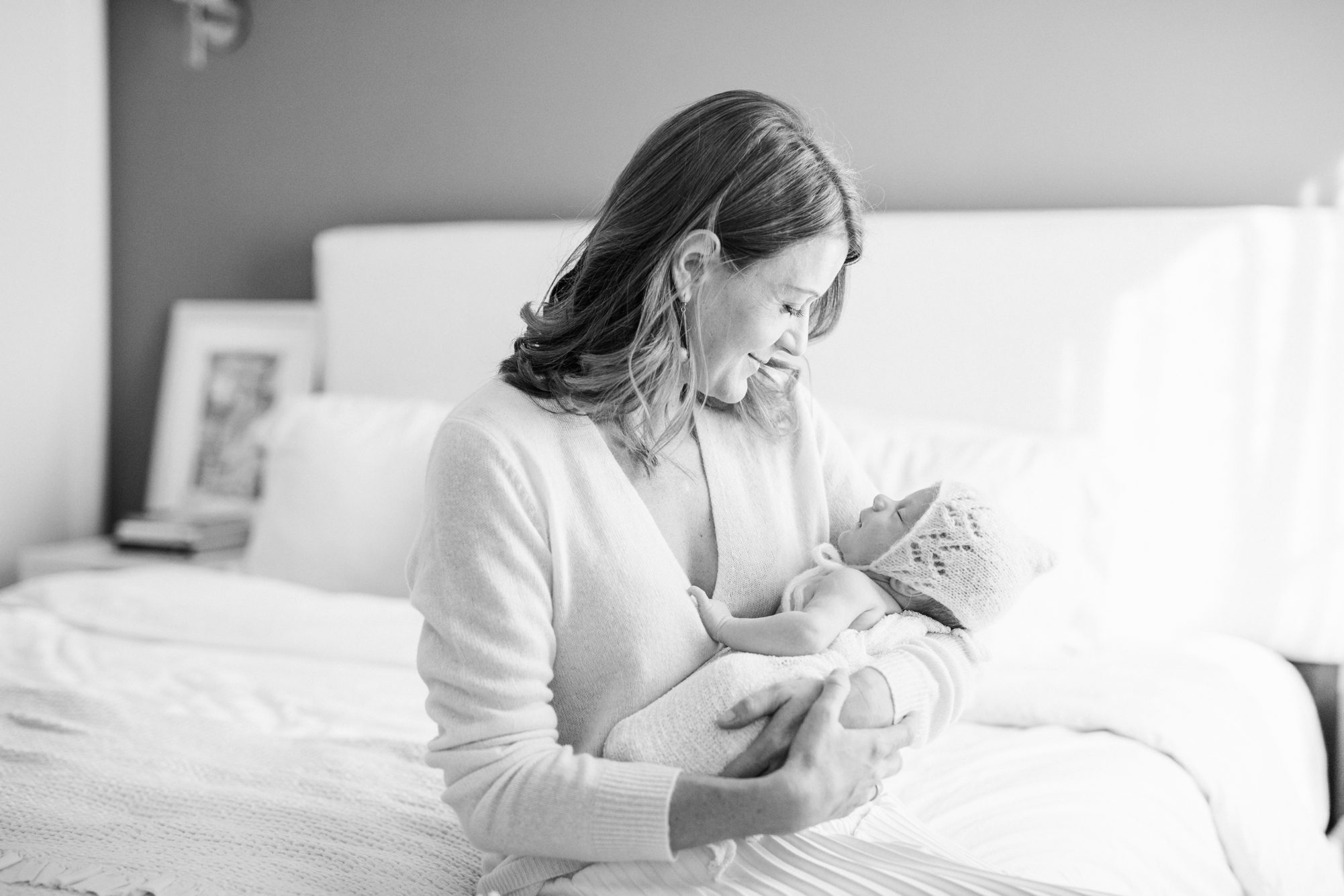 Black and white image of mom holding baby while sitting with him on edge of bed. Photo by LRG Portraits
