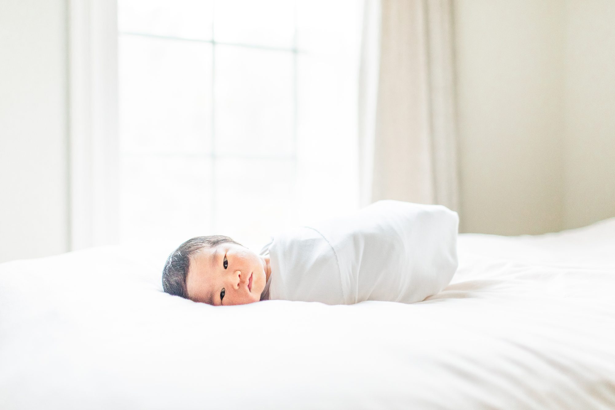 Baby in white swaddle on bed during in-home newborn session. Photo by LRG Portraits.