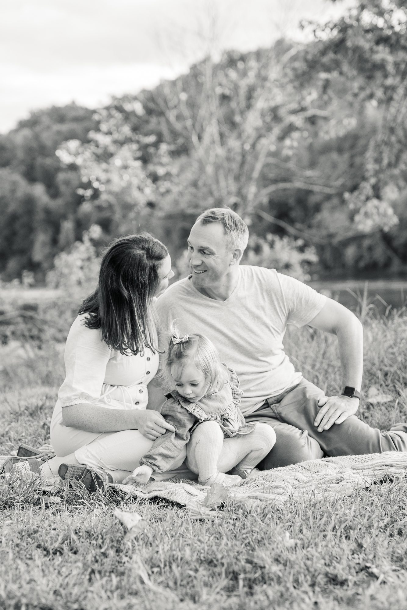 Black and white image of parents smiling at each other while toddler plays. Photo by LRG Portraits.