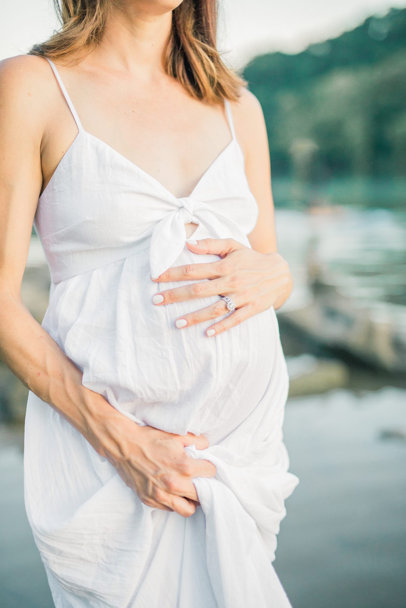 Cropped image of mom holding dress and resting hand on baby bump by Potomac River. Photo by Washington DC maternity photographer, LRG Portraits.