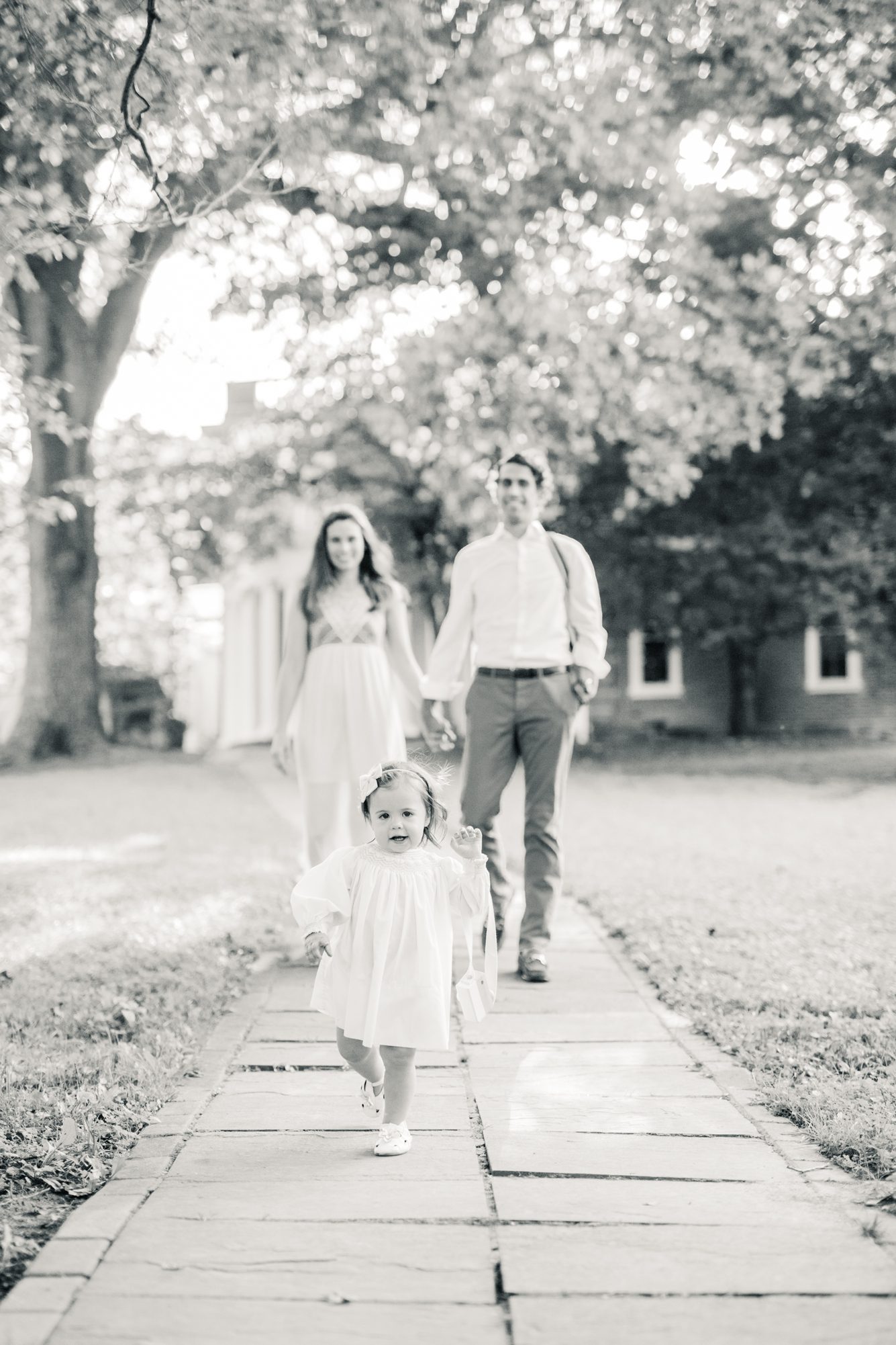 Black and white image of family walking together with toddler running ahead. Photo by LRG Portraits.