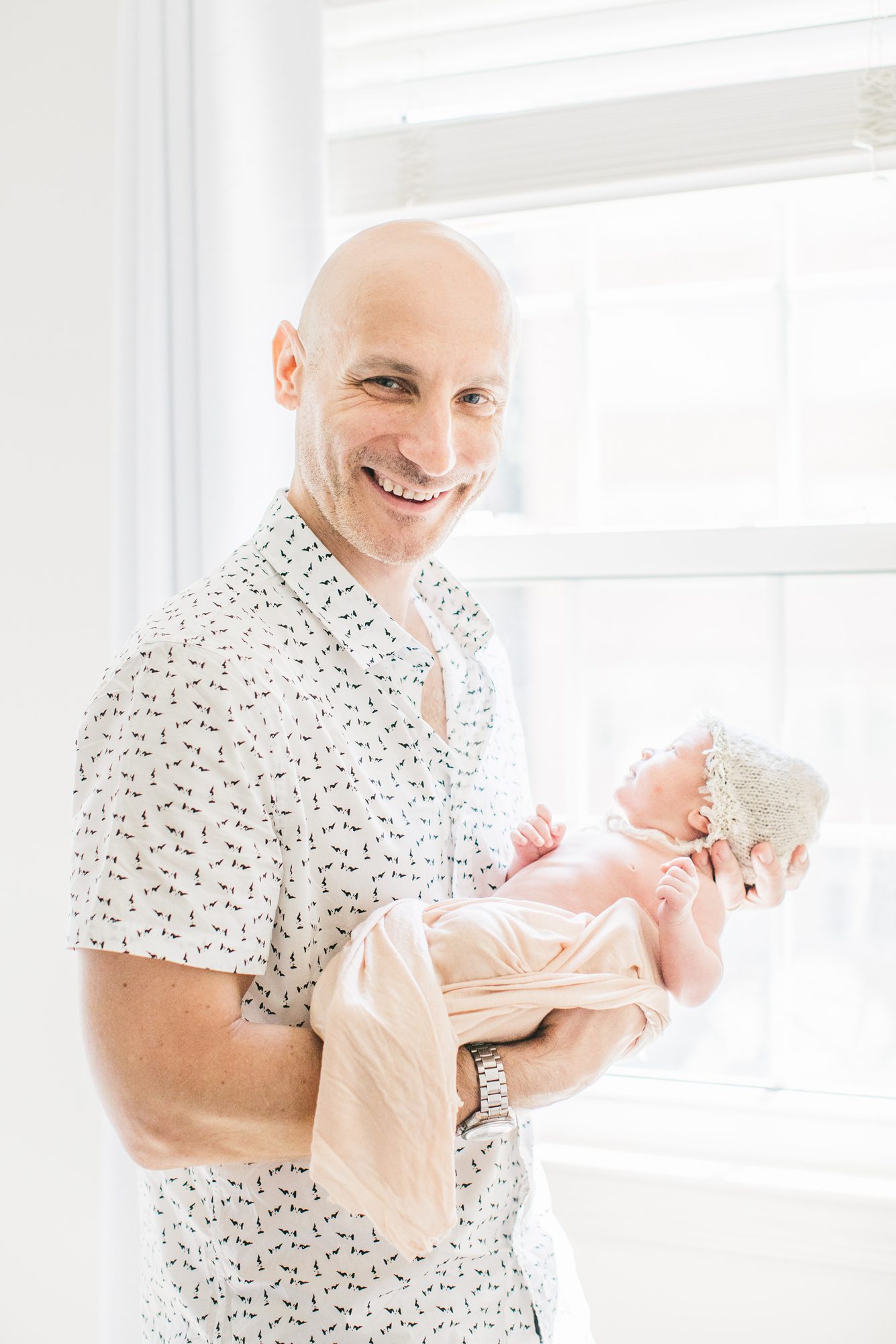 Dad smiling at camera while holding baby near window light. Photo by LRG Portraits.